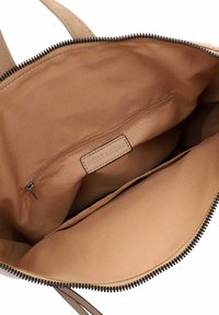 Interno di una grande borsa beige con tessuto liscio, chiusura a zip, tasca interna e etichetta del marchio. Cerniera nera testurizzata lungo il bordo.