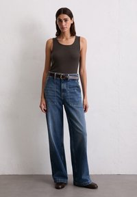 Frau trägt dunkel ärmelloses Oberteil, hoch taillierte, weit geschnittene blaue Jeans mit großen Vorderentaschen, braunen Gürtel und braune Schuhe, steht vor weißer Wand.