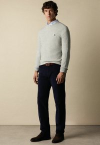 Lichtgrijze wollen trui met een ronde hals, donkerblauwe corduroy broek en bruine leren schoenen. Subtiel logo op de trui.