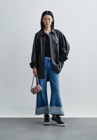 Frau trägt übergroße schwarze Lederjacke, schwarzes Oberteil, weit umgeschlagene blaue Jeans, schwarze Plateausneaker, hält kleine rosa Handtasche.