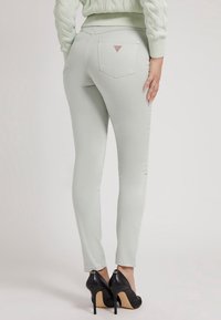 Kvinna i ljusgrå slim-fit jeans och svarta högklackade skor, stående med ena handen vilande på höften, vit bakgrund.