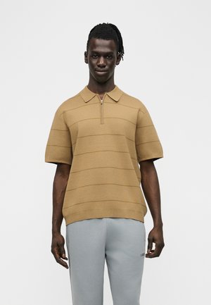 Άνδρας με σκοτεινό δέρμα και dreadlocks, που φοράει κοντομάνικο πουκάμισο τύπου πόλο με φερμουάρ σε χρώμα καμηλό και ανοιχτόγριμο παντελόνι, στέκεται μπροστά σε απλό φόντο.