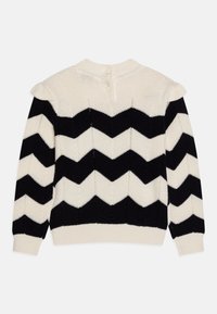 Pull en tricot crème et noir avec un motif en zigzag, col rond, et détails en boutons au dos avec de courtes accentuations en volants en tissu.