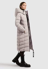 Dlhý, svetlo šedý puffer kabát s kapucňou, obsahujúci diamantový prešívaný vzor, bočné vrecká a zips. Nosený s čiernymi robustnými čižmami.