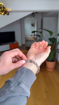 Brățară metalică cu zale interconectate și închizătoare cu lacăt, prezentată pe un încheietura mâinii. Cămașă gri cu mâneci lungi vizibilă. Mediu interior.