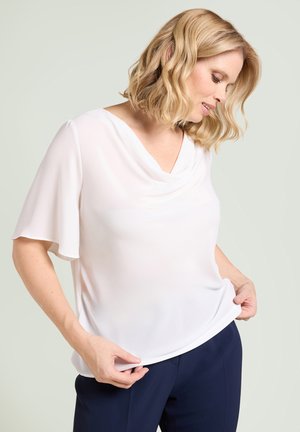 Femme blonde portant un chemisier blanc à manches courtes avec un col drapé, ajustant l'ourlet, et un pantalon bleu marine sur un fond clair uni.