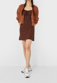 Bruine geribbelde cardigan, zwarte jurk met luipaardprint en beige/witte sneakers. De jurk heeft verstelbare bandjes, de cardigan is voorzien van knopen.