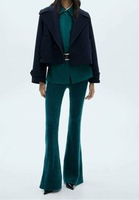 Giacca doppiopetto corta blu navy, camicia teal sovrapposta, e pantaloni teal svasati in un tessuto morbido con scarpe nere a punta.