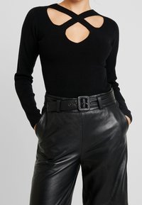 Svart stickad topp med korsade skärningar, ihopparad med högmidjade svarta läderbyxor som har ett brett bälte och sidofickor.