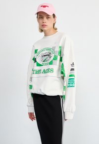 White sweatshirt with green and gray checkered pattern, featuring a circular graphic and text. Paired with a black ribbed skirt and pink cap.