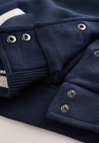 Tissu bleu marine avec boutons-pression argentés et bords côtelés, montrant des détails en gros plan de la fermeture et de la couture d'un vêtement.