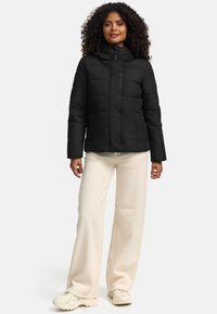 Black puffer jacket with a high collar, quilted design, front zip closure, and two side pockets, worn over cream wide-leg pants.