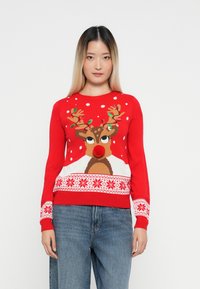 Fiatal nő piros karácsonyi pulóvert visel, amelyen díszített rénszarvas és hópehely minták találhatók, kék farmernadrággal párosítva.