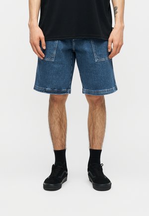 Uomo che indossa pantaloncini di denim blu con tasche anteriori, calze nere e scarpe da ginnastica nere, in piedi contro uno sfondo chiaro e uniforme.
