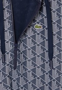 Temno modra zip-up jakna z geometrijskim vzorcem v belem. Ima označen zadrgo in majhen zeleni krokodilov logotip na prsih.