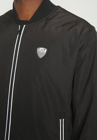 Svart bomberjakke laget av nylon, med glidelås foran, hvite glidelåsdetaljer, ribbet krage og et lite logo-patch på venstre bryst.