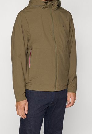 Homme portant une veste à capuche vert olive avec une fermeture éclair avant et un jean bleu foncé, debout devant un fond clair uni.