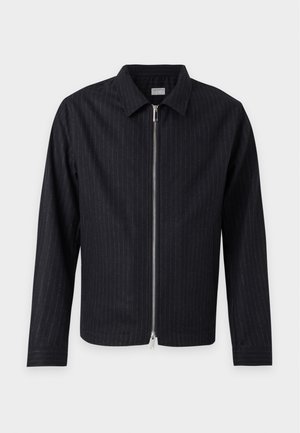 Ung mand stående iført en sort lynlåstrøje, beige poloshirt, brede beige bukser, sorte sko, med den ene hånd i lommen mod en ensfarvet baggrund.
