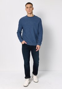 Suéter azul de punto con cuello redondo, combinado con jeans oscuros y zapatillas blancas. La textura parece suave y el ajuste es informal.