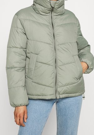 Personne portant une doudoune matelassée vert clair avec fermeture éclair et poches à l'avant, associée à un jean bleu clair, debout devant un fond neutre.
