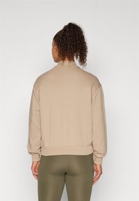 Pullover felpa color sabbia con collo alto, vestibilità oversize e polsini a costine. Texture liscia, indossata con leggings verde oliva.