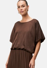 Blusa marrone, leggera e fluida, con scollatura ampia e maniche corte e larghe, caratterizzata da una texture liscia e una vestibilità comoda.