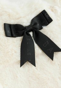 Kreminė sintetinio kailio medžiaga su juodu satino kaspinu su įspaustomis raidėmis, surištu centre. Kaspino galiukai yra smailūs ir ryškiai kyšančios.