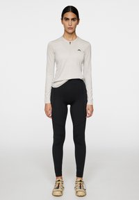 Ljust beige långärmad tröja med dragkedja och logotyp, i kombination med högmidjade svarta leggings. Modellen bär beige skor med flerfärgade accenter.