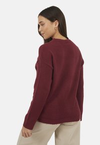 Pull en maille bordeaux à coupe décontractée, avec poignets côtelés et encolure arrondie, offrant une texture douce et un ourlet arrière légèrement plus long.