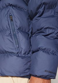 Casaco puffer azul-marinho com bolso no peito com zipper, design acolchoado e punhos canelados; o material parece suave com leve brilho.