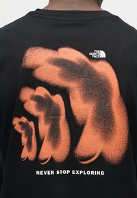 Πλάτη μαύρου T-shirt με πορτοκαλί αφηρημένα σχήματα χεριών, λογότυπο The North Face και το κείμενο "NEVER STOP EXPLORING" σε λευκό χρώμα.
