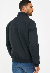 Svart bomberjacka med ståkrage, texturerad tyg och ribbade ärmslut. Bärs med blå jeans och visar en minimalistisk design.