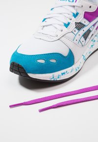 White and turquoise athletic shoe with mesh and suede materials, featuring purple accents and textured sole. Attached are two purple laces.