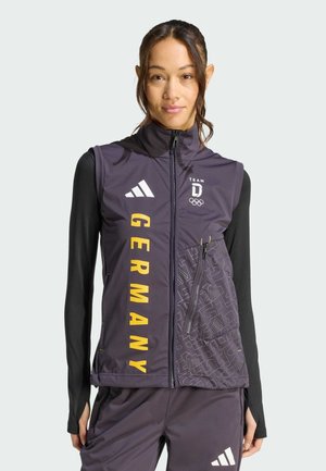 Woman wearing black long sleeves under a dark gray Team Germany vest with yellow text and Adidas logo, standing against a plain background.