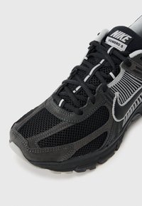 Μαύρο αθλητικό παπούτσι με υλικά από δίχτυ και σουέντ, με γκρι λογότυπο Nike, δομημένο σχέδιο και λευκές λεπτομέρειες στα κορδόνια και στις πλευρές.