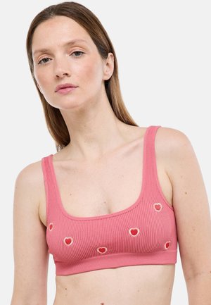 Femme aux cheveux bruns raides portant un crop top côtelé rose avec des cœurs brodés rouges et blancs sur un fond uni.