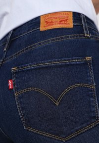 Jeans de mezclilla azul oscuro con un parche de cuero en la parte trasera que dice "Levi Strauss & Co." y una etiqueta roja en el bolsillo trasero con un diseño curvado.