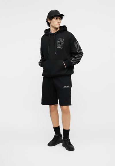 Jauns vīrietis tērpies melnā Off-White džemperī ar kapuci, melnās šortās, melnā cepurē, melnās zeķēs un melnos sporta apavos, stāvot pret vienkāršu fonu.