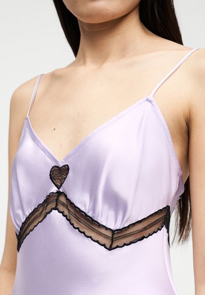 Γυναίκα που φοράει λιλά σατέν καμιζόλ με λεπτούς ιμάντες, με μαύρες δαντέλες σε σχήμα καρδιάς και ζιγκ-ζαγκ στο στήθος και την κοιλιά.