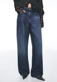 Leveälahkeiset siniset farkut, joissa haalistettu viimeistely, kiristysnyörivyötärö, viisi taskua ja yksityiskohtaiset ompelut.