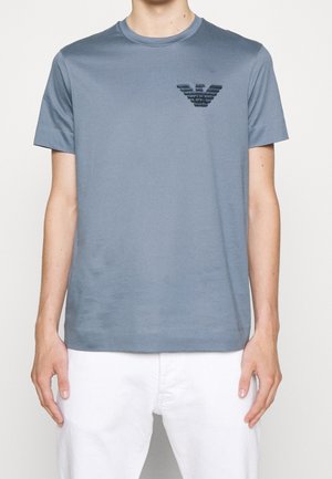 T-shirt gris moyen à manches courtes avec un petit emblème noir sur le côté gauche de la poitrine, porté avec un pantalon blanc.