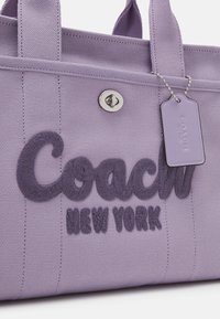 Violetti kangaskassi, jossa kohokuvioitu "Coach New York" -kirjailu, hopeiset metalliosat ja ketjulla kiinnitetty tunniste.