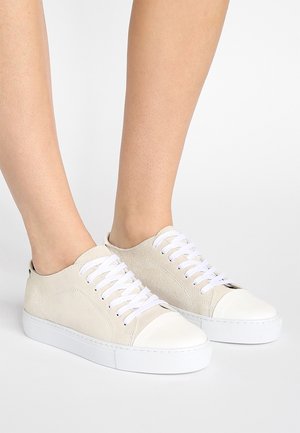 Baskets basses en daim beige avec embouts et semelles en caoutchouc blanc, portées pieds nus sur un fond clair uni.