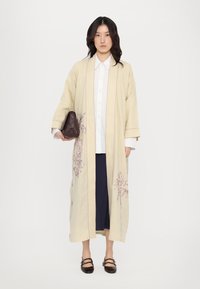 Dlhý béžový kimono s ružovou výšivkou, širokými rukávmi a detailným kvetinovým vzorom. V kombinácii s bielou košeľou a tmavými nohavicami, s hnedou taškou v ruke.
