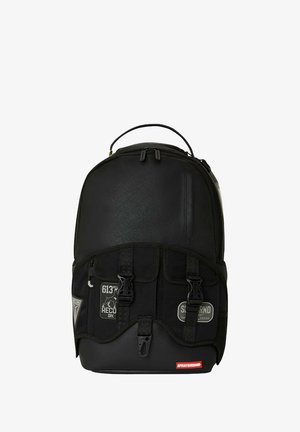 Mochila negra con tela texturizada, correas ajustables, dos hebillas en la parte frontal y parches con logotipo. Compartimento principal con cremallera y bolsillo lateral.