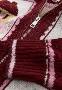Roter und pink gemusterter Strickpullover mit Reißverschluss; verfügt über eine weiche Textur, gerippte Bündchen und kontrastierende Streifen.