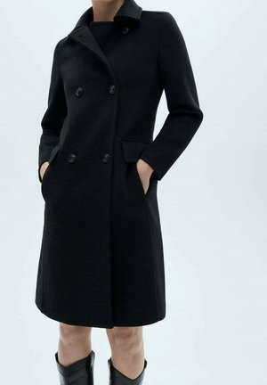 Donna che indossa un cappotto nero doppiopetto fino al ginocchio con tasche laterali e stivali neri, in piedi contro uno sfondo chiaro semplice.