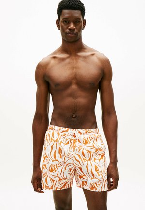 Sportlicher Mann, der nach vorne blickt, trägt orange-weiße Badehose mit tropischem Muster, oben ohne vor einfachem Hintergrund.