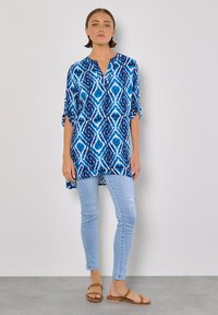 Blautönige Tunika mit lockerer Passform, V-Ausschnitt und umgeschlagenen Ärmeln. Kombiniert mit hellblauen Skinny-Jeans und flachen Sandalen.