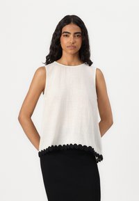 Haut sans manches crème en tissu texturé, avec un ourlet festonné noir. Assorti à une jupe noire unie.